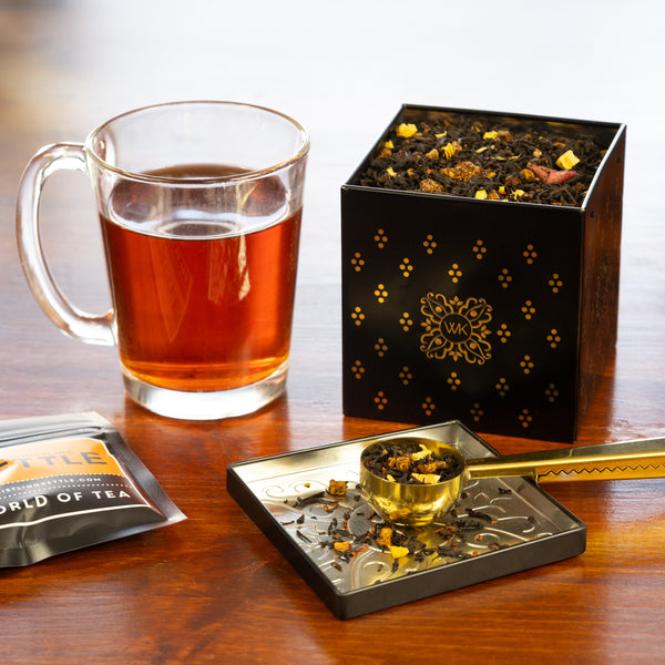 A tea tin of Caramel Apple Bourbon tea leaves sits on a table next to a cup of brewed tea