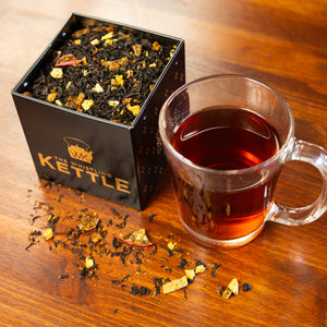 A tea tin of Caramel Apple Bourbon tea leaves sits on a table next to a cup of brewed tea