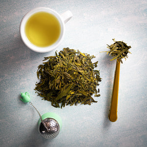 Cup of Dragonwell tea next to a mound of loose leaf Dragonwell and other teaware.