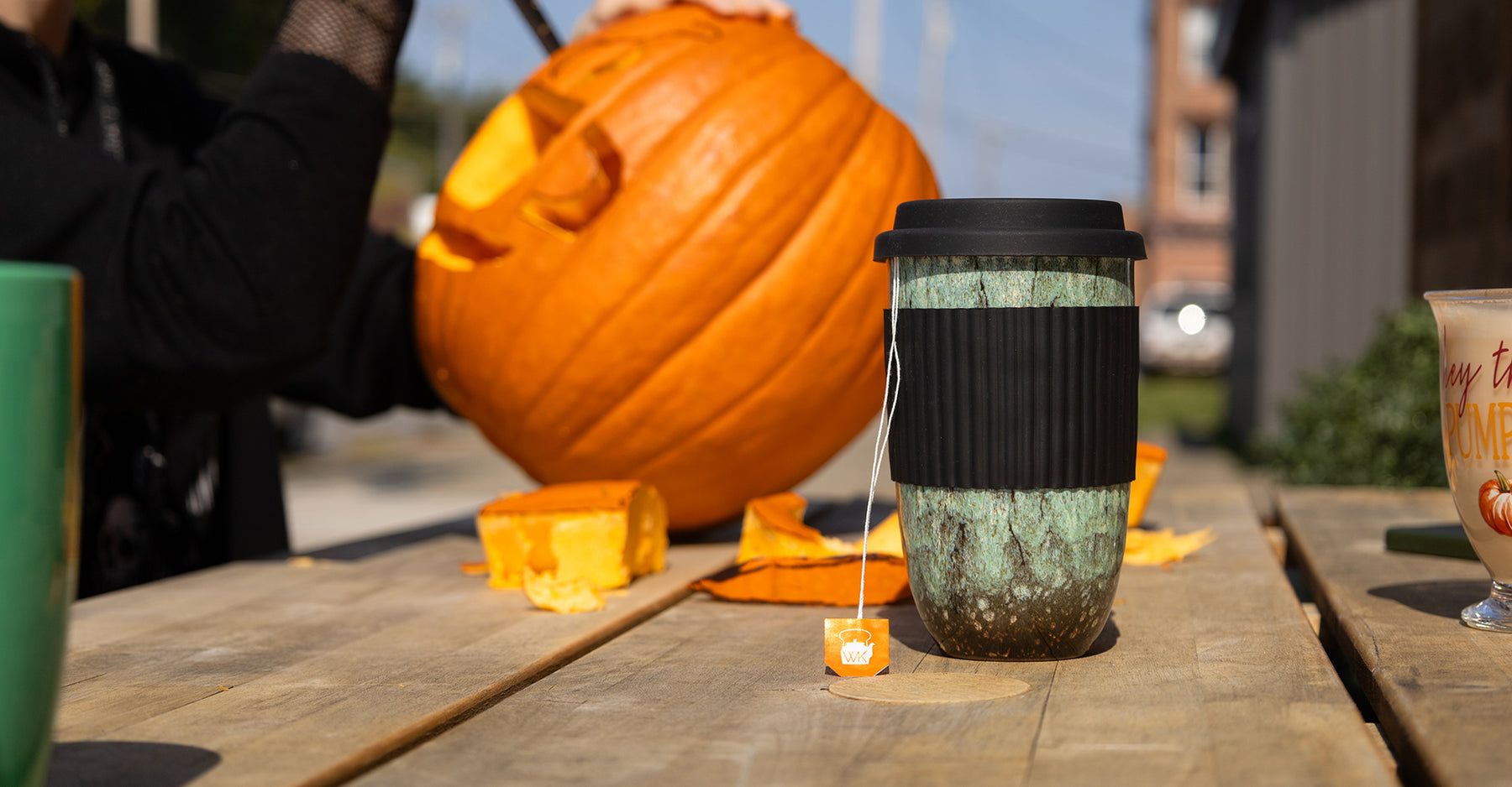 A travel cup of tea sits on a picnic table while someone carves a pumpkin in the background