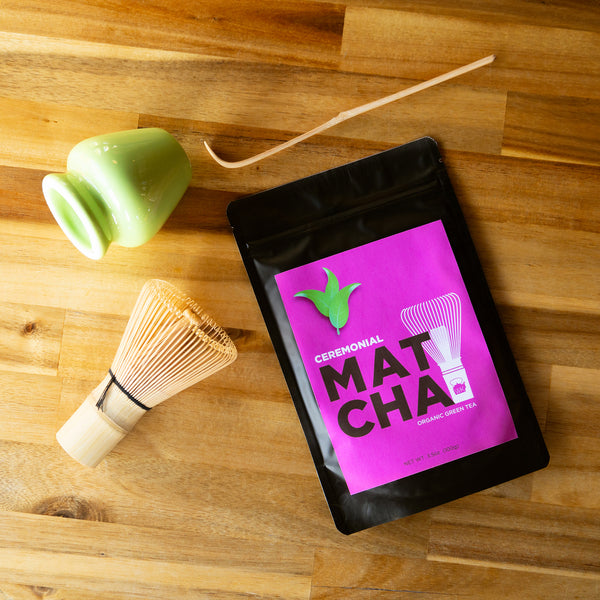 A bag of ceremonial matcha, a whisk, a whisk stand, and a matcha spoon lay on a wooden table.